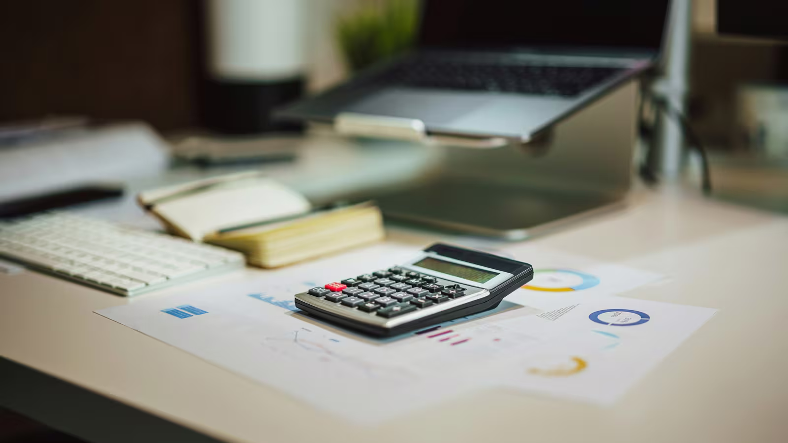 Calculatrice posée sur un bureau à côté d'un ordinateur portable et de graphiques budgétaires imprimés (photo Jakub Żerdzicki / Unsplash)