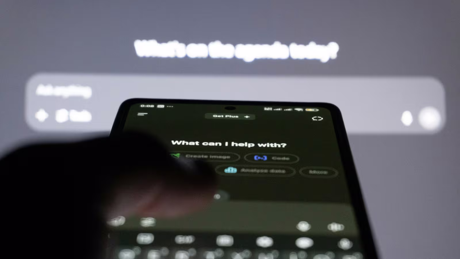 Une main tient un smartphone ouvert sur l'application ChatGPT, en arrière-plan un écran affiche lui aussi une interface d'IA conversationnelle (photo Zulfugar Karimov / Unsplash)